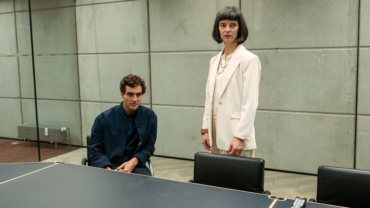 Die Gründer von Prometheus Dr. Oskar Berger und Ehefrau Viola sitzen bzw. stehen in einem nüchternen Besprechungsraum.