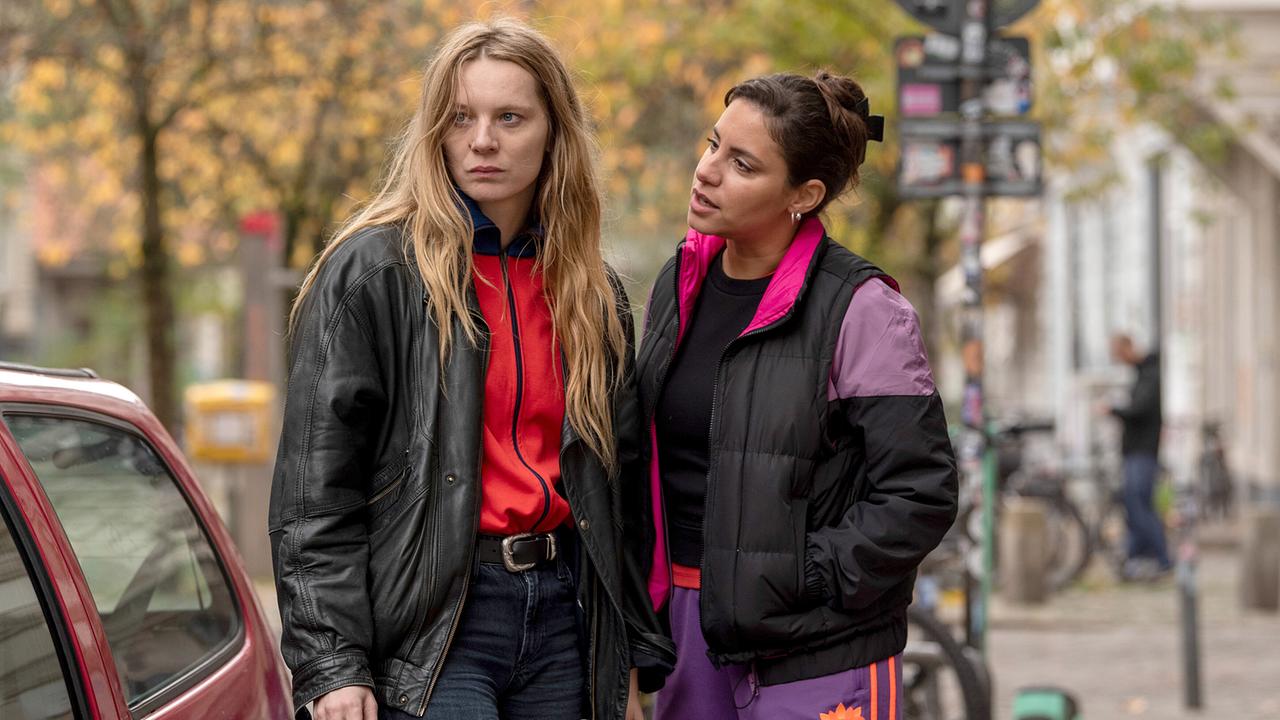 Anna Nowak und Mila Sahin stehen neben einem roten Auto auf einer herbstlichen Straße in Kiel.