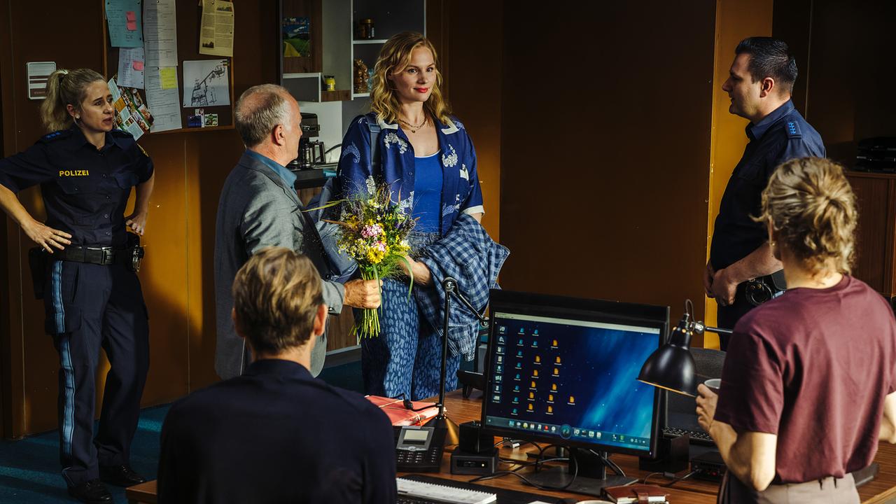 Dr. Mirko Kaiser mit einem Blumenstrauss in der Hand stellt den Kollegen die neue Kollegin Emmy Rathgeber im Polizeibüro vor.