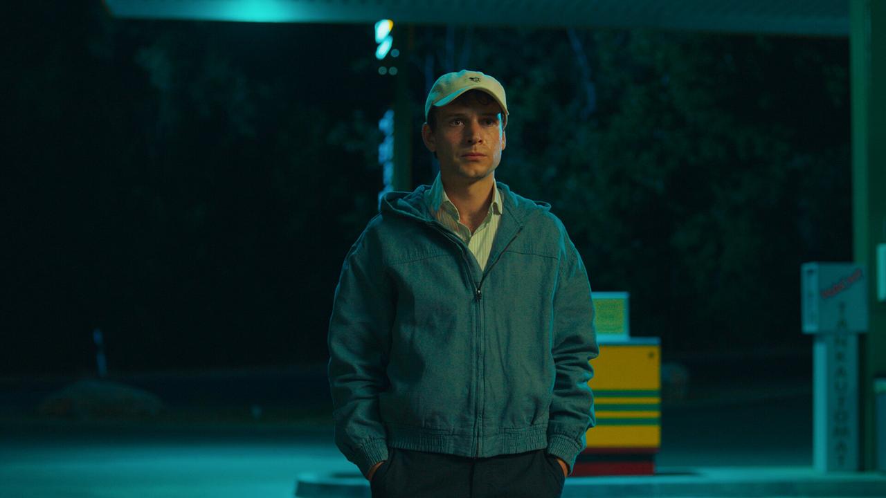 Toni mit Kappe steht nachts allein an einer beleuchteten Tankstelle.