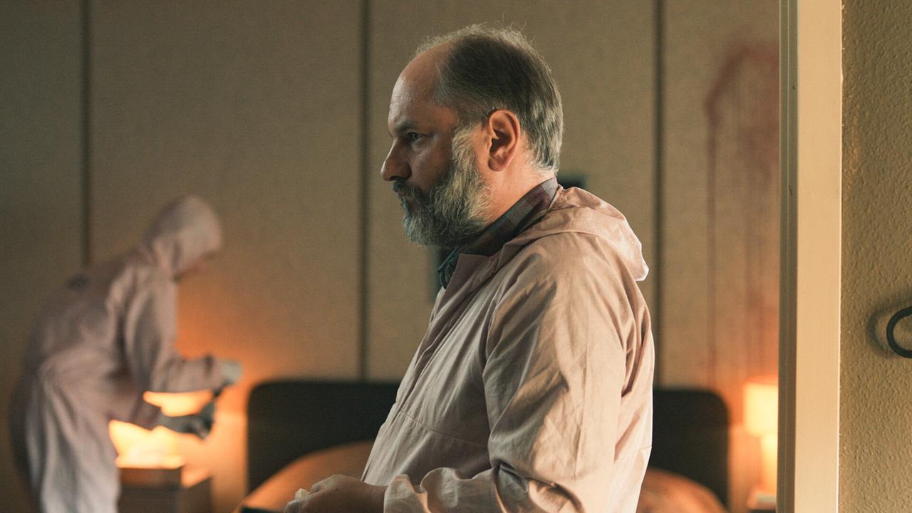 Michael Schatz in Schutzkleidung steht in einem Raum bei der Arbeit am Tatort, während im Hintergrund eine Person mit einer Kamera arbeitet.