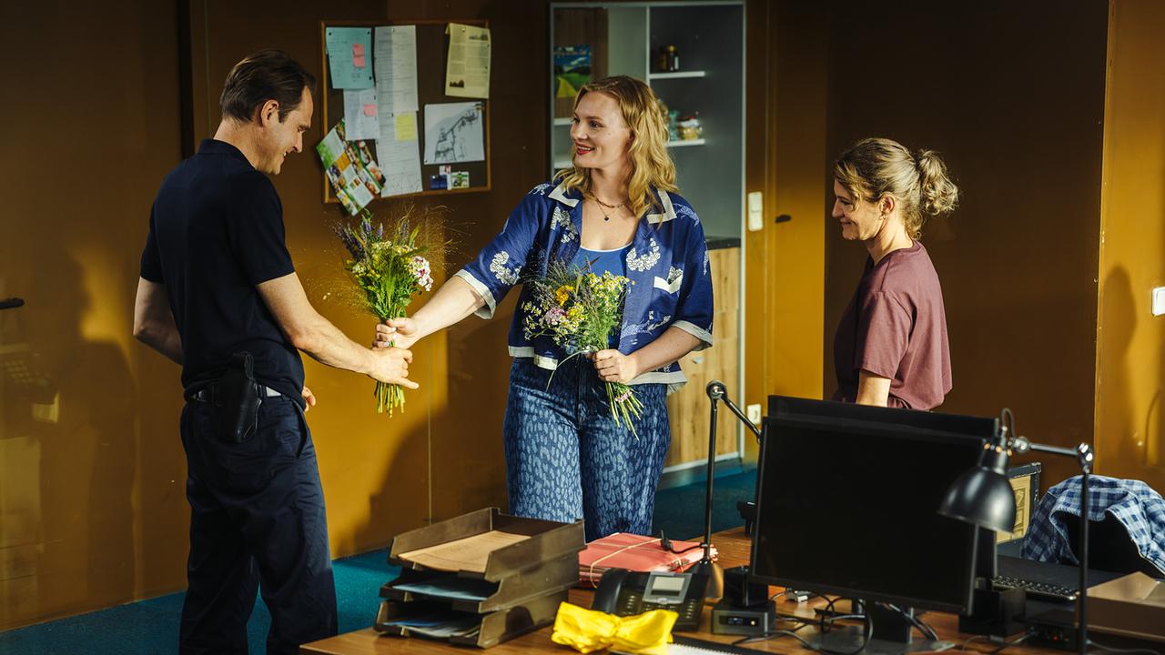 Da Felix Voss Geburtstag hat, reicht ihm Emmy Rathgeber im Kommissariat die Hälfte ihres Blumenstrausses, den sie zur Begrüßung erhalten hat. Darüber muss Wanda Goldwasser  schmunzeln.