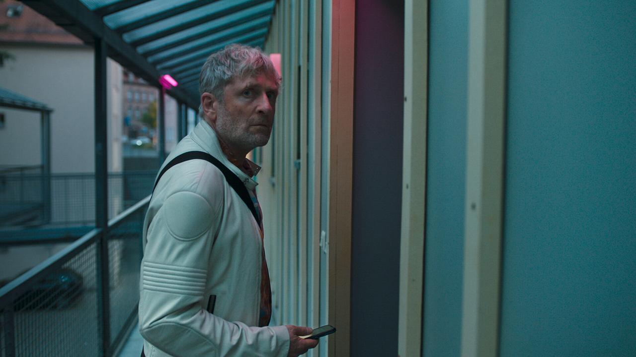 Thorsten Widner in heller Jacke steht mit seinem Handy vor einer Wohnungstür in einem Außengang und fühlt sich beobachtet.