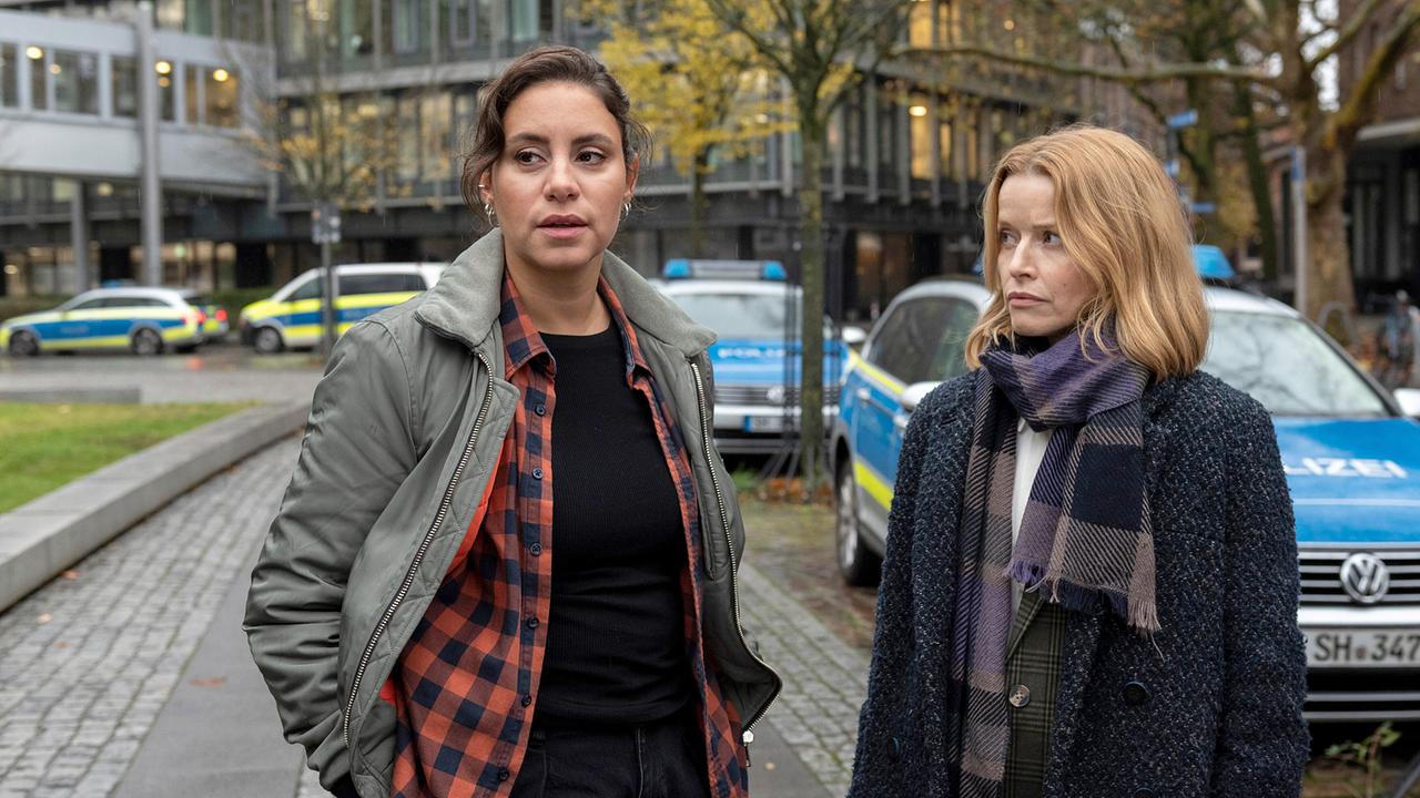 Mila und Kriminalpsychologin Elli Krieger stehen vor Polizeiwagen auf einer herbstlichen Straße und blicken angespannt in unterschiedliche Richtungen.