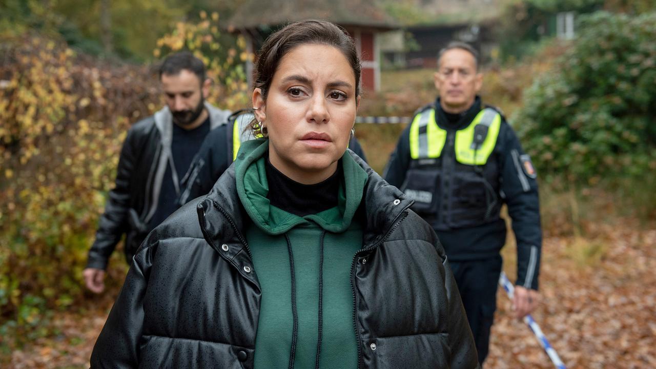 Die besorgt wirkende Mila geht im Regen voraus, hinter ihr folgen ein Mann und ein Polizist auf einem herbstlichen Weg zum Haus am See.