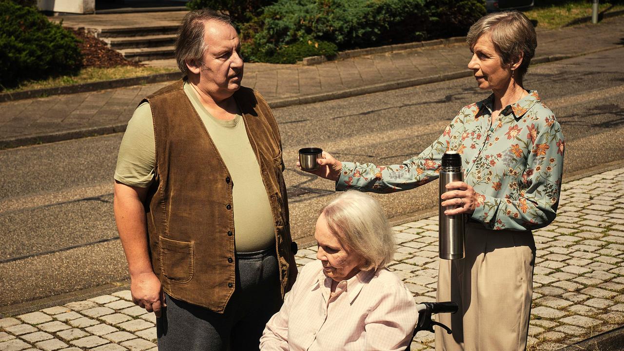 Anna Stieblich bietet am Gehsteig Wolf Bachofner aus einer Thermoskanne ein Getränk an. Eine Frau im Rollstuhl sitzt vor ihnen.