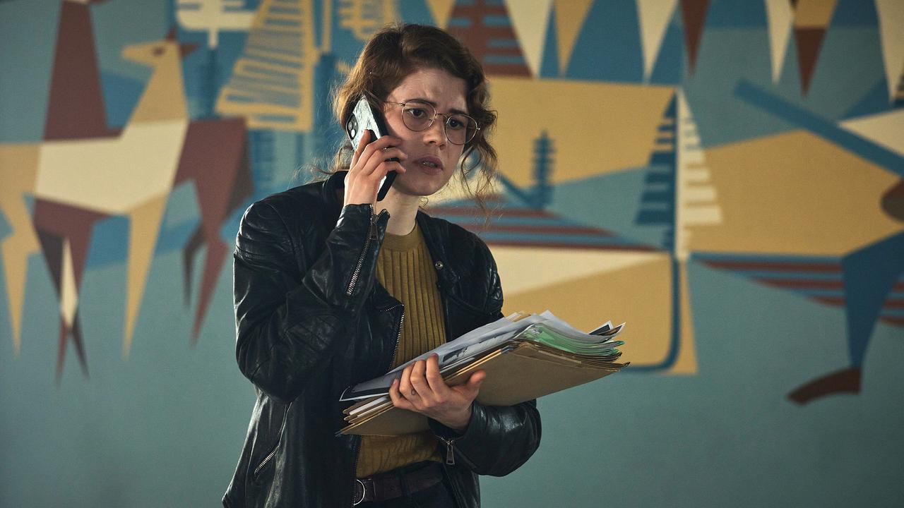 Luise Aschenbrenner telefoniert und hält einen Stapel Akten, vor bunter Wandmalerei.