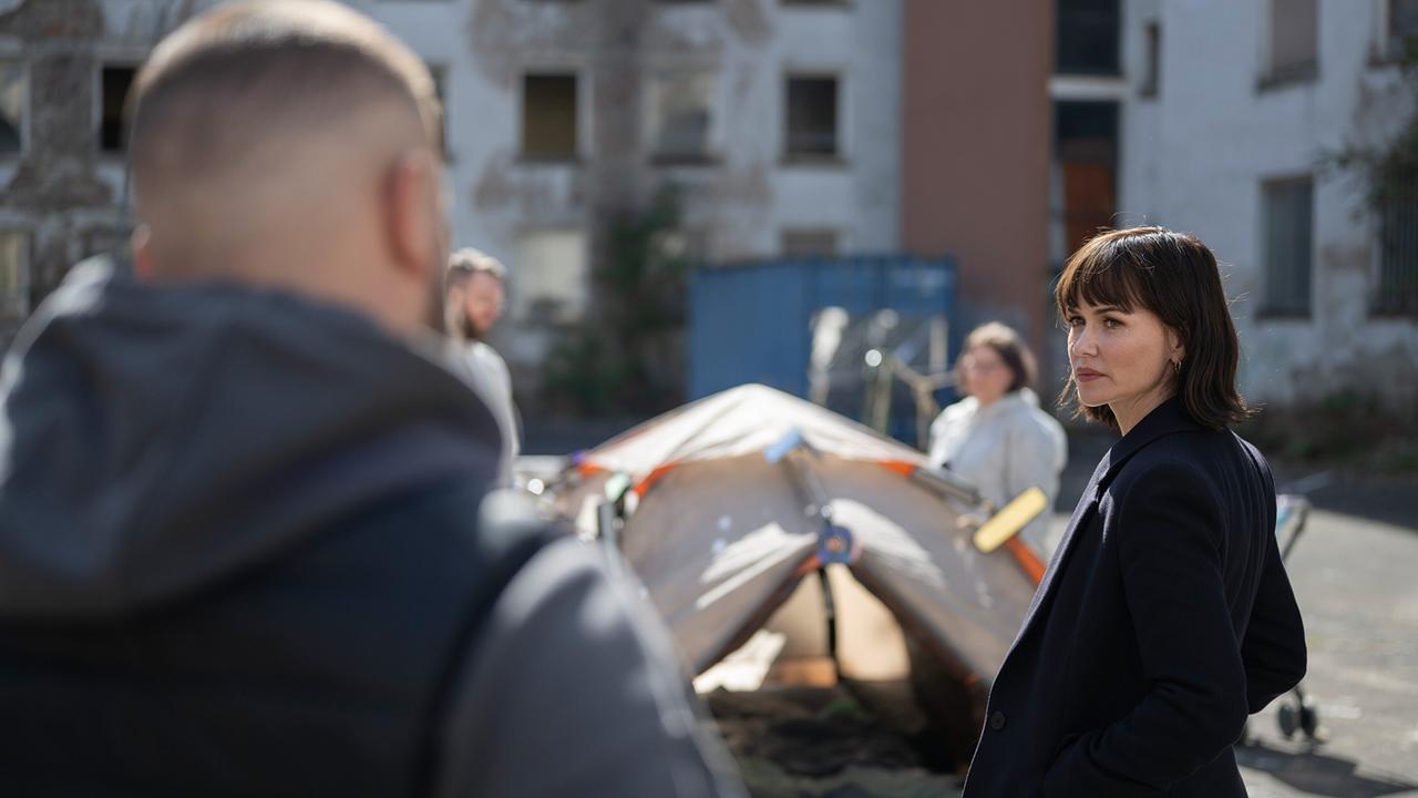 © ORF/HR/Sommerhaus/Daniel Dornhöfer Edin Hasanivic und Melika Foroutan schauen sich in einem Hof vor einem Zelt mit weiteren Menschen in weißen Schutzanzügen an.