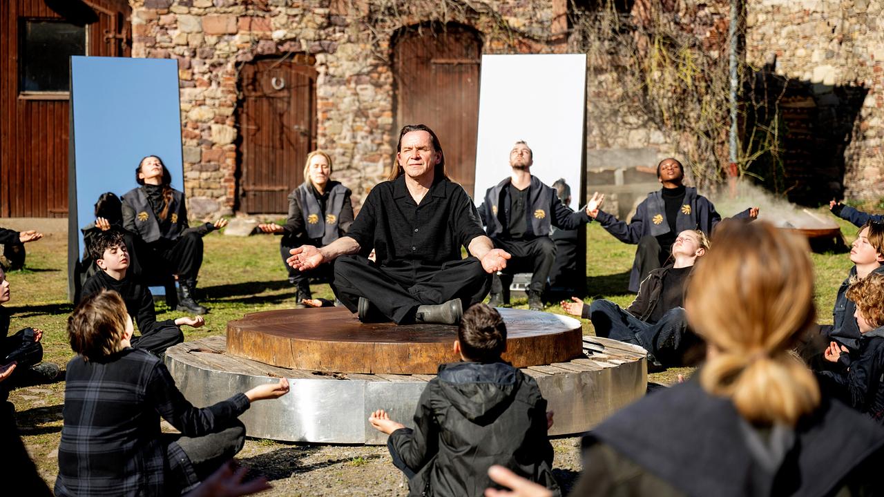 © ORF/HR/Sommerhaus/Daniel Dornhöfer Mitglieder der Lichtsekte sitzen mit geöffneten Armen und Kopf nach oben gerichtet im Freien im Kreis, während eine Person auf einer runden Plattform meditiert.