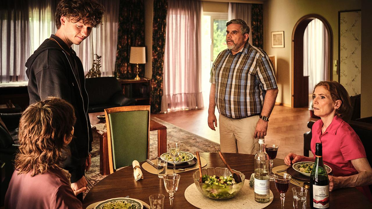 In einem Esszimmer sitzen und stehen Mikes Eltern und Mike und Nisha um einen gedeckten Tisch mit Salat und Wein.