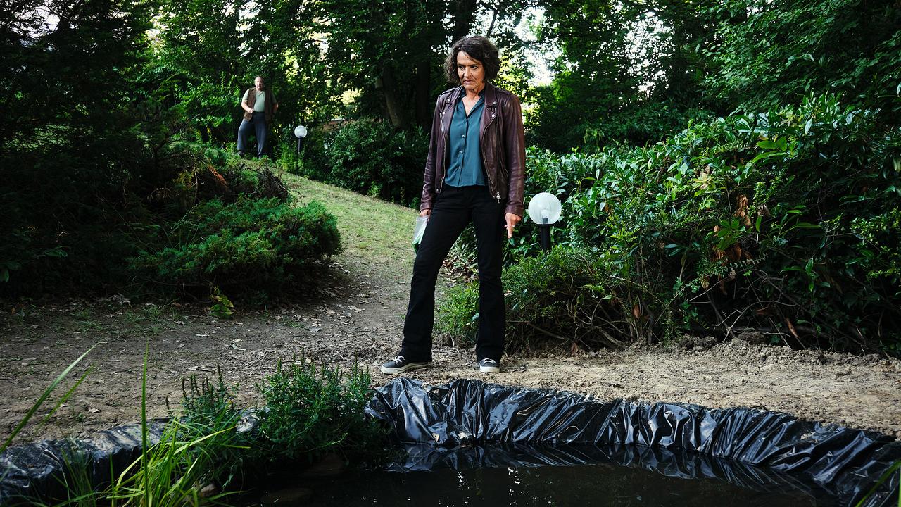  Lena Odenthal in Lederjacke steht am Rand eines frisch angelegten Gartenteichs, während im Hintergrund Erwin Ramthor auf sie blickt.