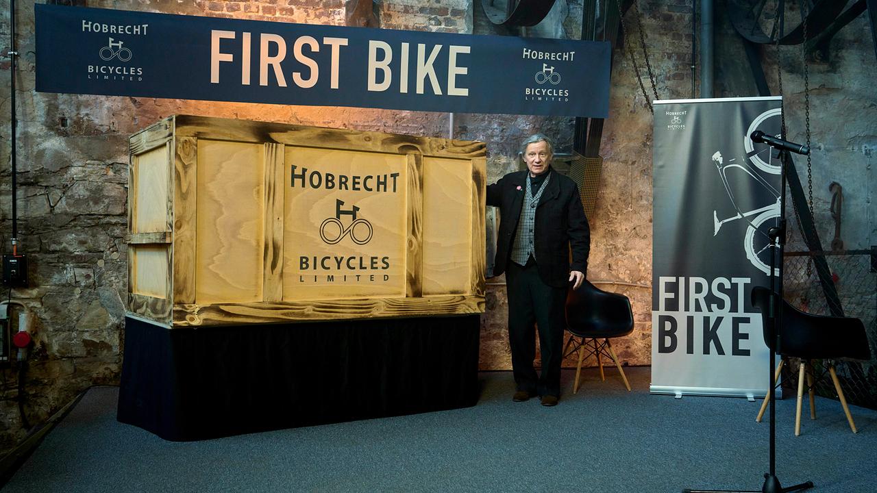 Der Patriarch der Fahrrad-Dynastie Kurt Hobrecht sen. steht neben einem großem Holzkasten, darüber und davor sind „First Bike“-Banner zu sehen, und präsentiert stolz das Highlight seines Lebenswerks