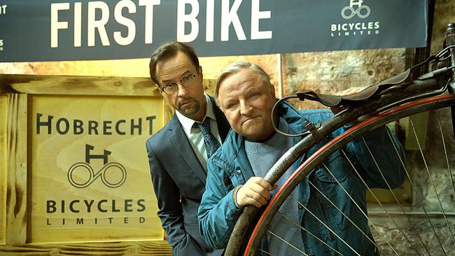 © ORF/WDR/Frank Dicks Prof. Karl Friedrich Boerne und Kommissar Frank Thiel schauen hinter einem Hochrad hervor, darüber ein „First Bike“-Banner.