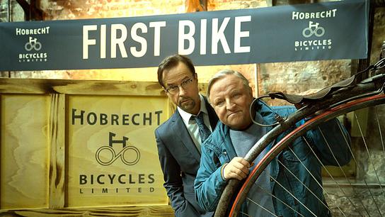 © ORF/WDR/Frank Dicks Prof. Karl Friedrich Boerne und Kommissar Frank Thiel schauen hinter einem Hochrad hervor, darüber ein „First Bike“-Banner.