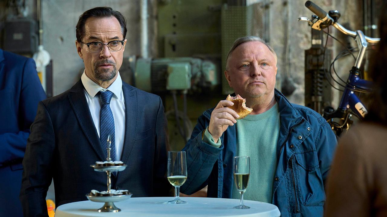 Prof. Karl Friedrich Boerne und Kommissar Frank Thiel stehen an Stehtisch mit Sektgläsern.  Thiel isst Gebäck.