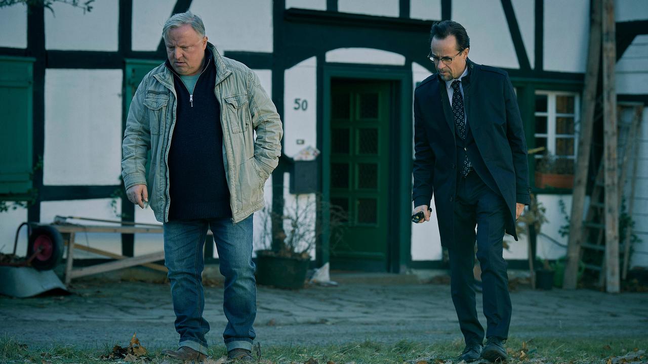 Kommissar Thiel und Prof. Boerne stehen vor einem Fachwerkhaus und schauen zu Boden.