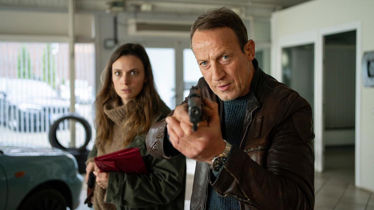 Falke zielt mit einer Schusswaffe in einem Autohaus, während Lynn de Baer hinter ihm steht und eine rote Mappe hält.
