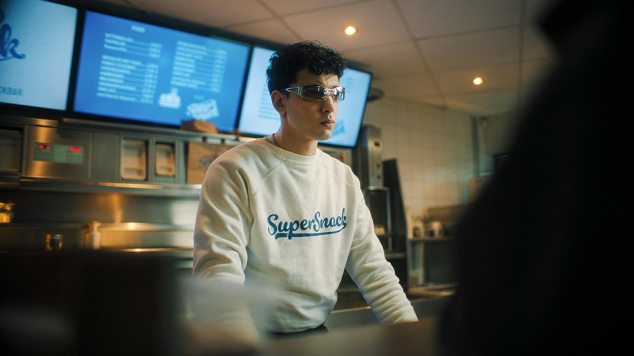 Imbiss-Aushilfe Sami mit einer Sonnenbrille steht hinter einer Imbiss-Theke mit „SuperSnack“-Logo auf dem Pullover.