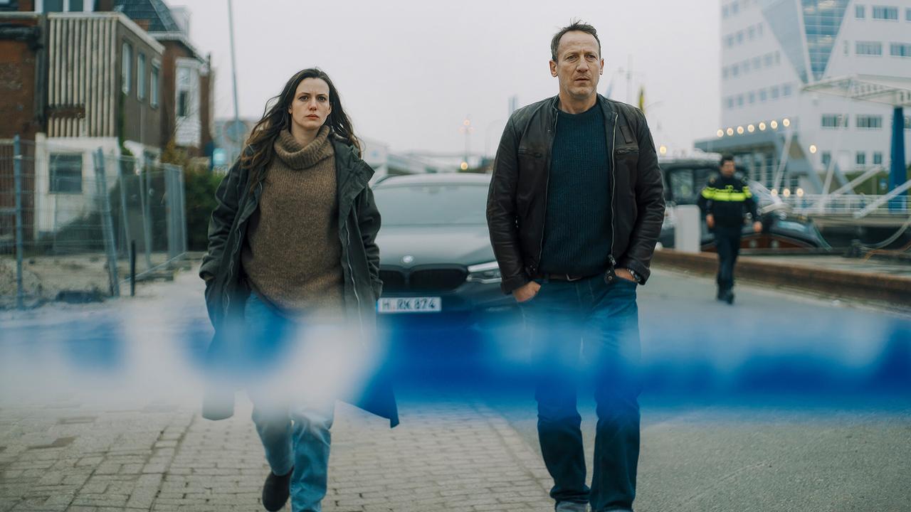 Werden zu einem Anschlag mit tödlichem Ausgang in Delfzijl gerufen: Lynn de Baer und Falke vor einem blauen Absperrband auf einer Uferstraße, während ein Polizist im Hintergrund zu sehen ist.