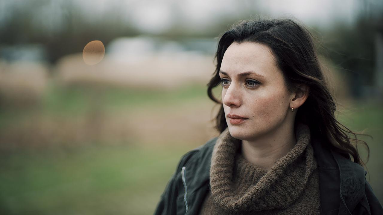 Kommissarin Lynn de Baer, Unterstützung der niederländischen Polizei für Falke, trägt einen braunen Wollpullover und steht im Freien, unscharfer Hintergrund.