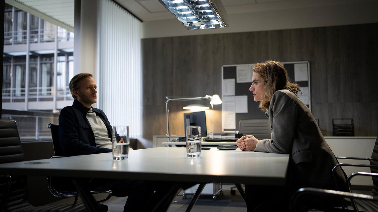 LKA-Chef Roland Krebcke sitzt Staatsanwältin Dr. Nadolny in einem modernen Büro an einem Tisch gegenüber, vor ihnen stehen Wassergläser.