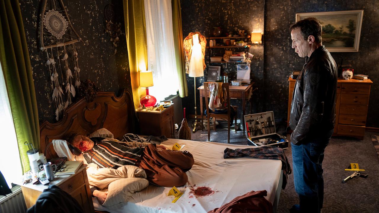  Falke steht in einem unordentlichen Schlafzimmer vor einer blutüberströmten Leiche im Bett, umgeben von Tatortmarkierungen.