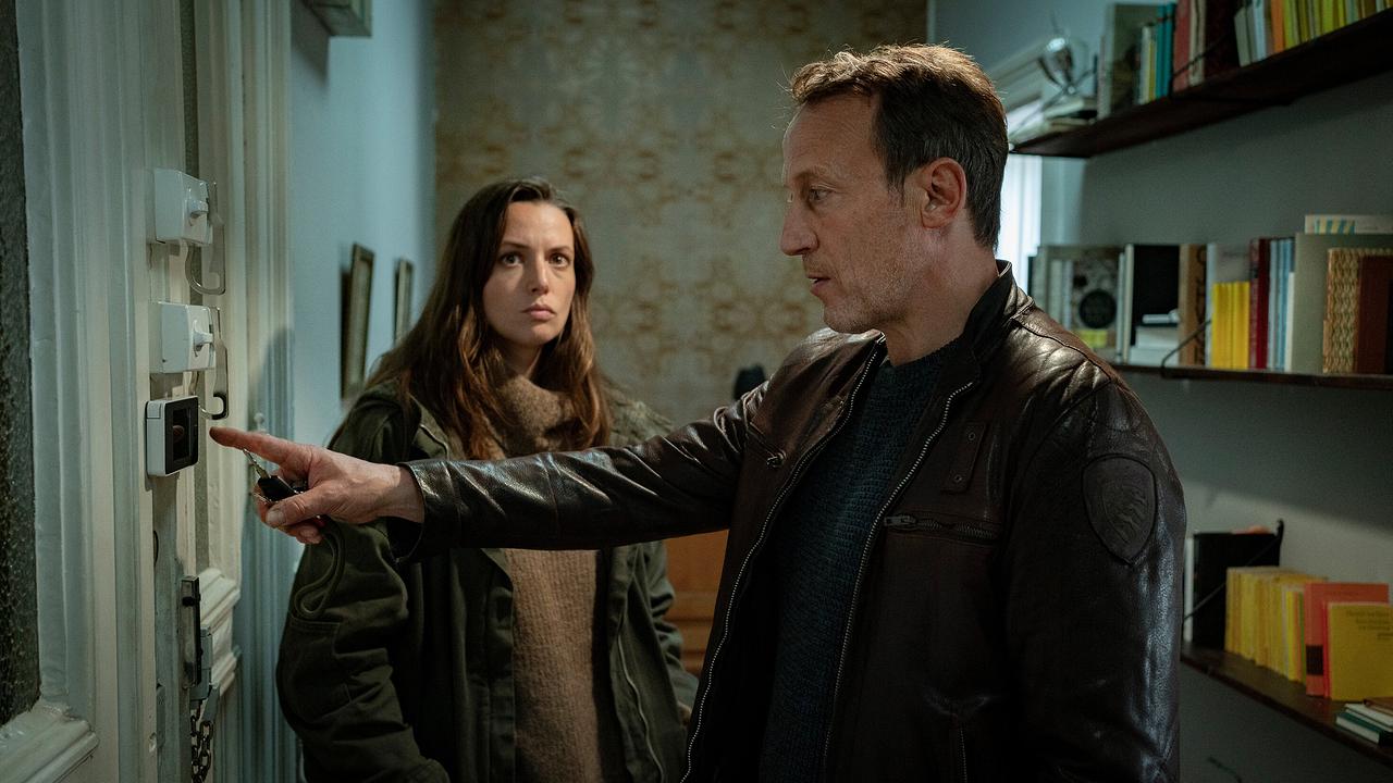 Lynn de Baer und Falke stehen nebeneinander in einem Innenraum und betrachten die Schlösser an der Eingangstür.