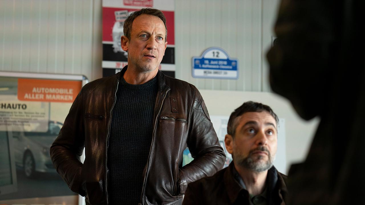 Falke in Lederjacke und Mario Schmitt im Büro eines Autohändlers schauen auf etwas vor ihnen.