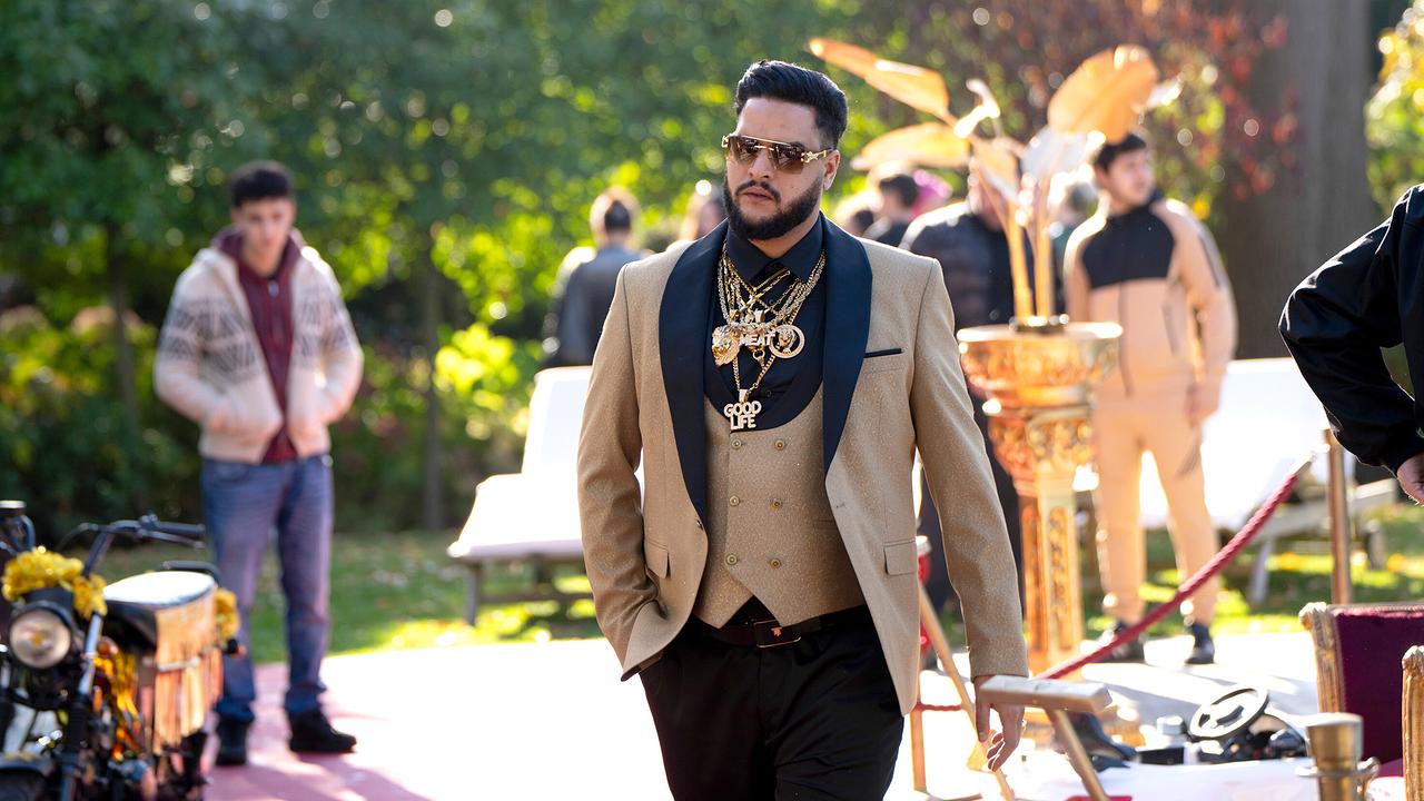 Karim Saidi in gold-schwartem Anzug mit auffälligen Halsketten geht über einen roten Teppich im Freien.