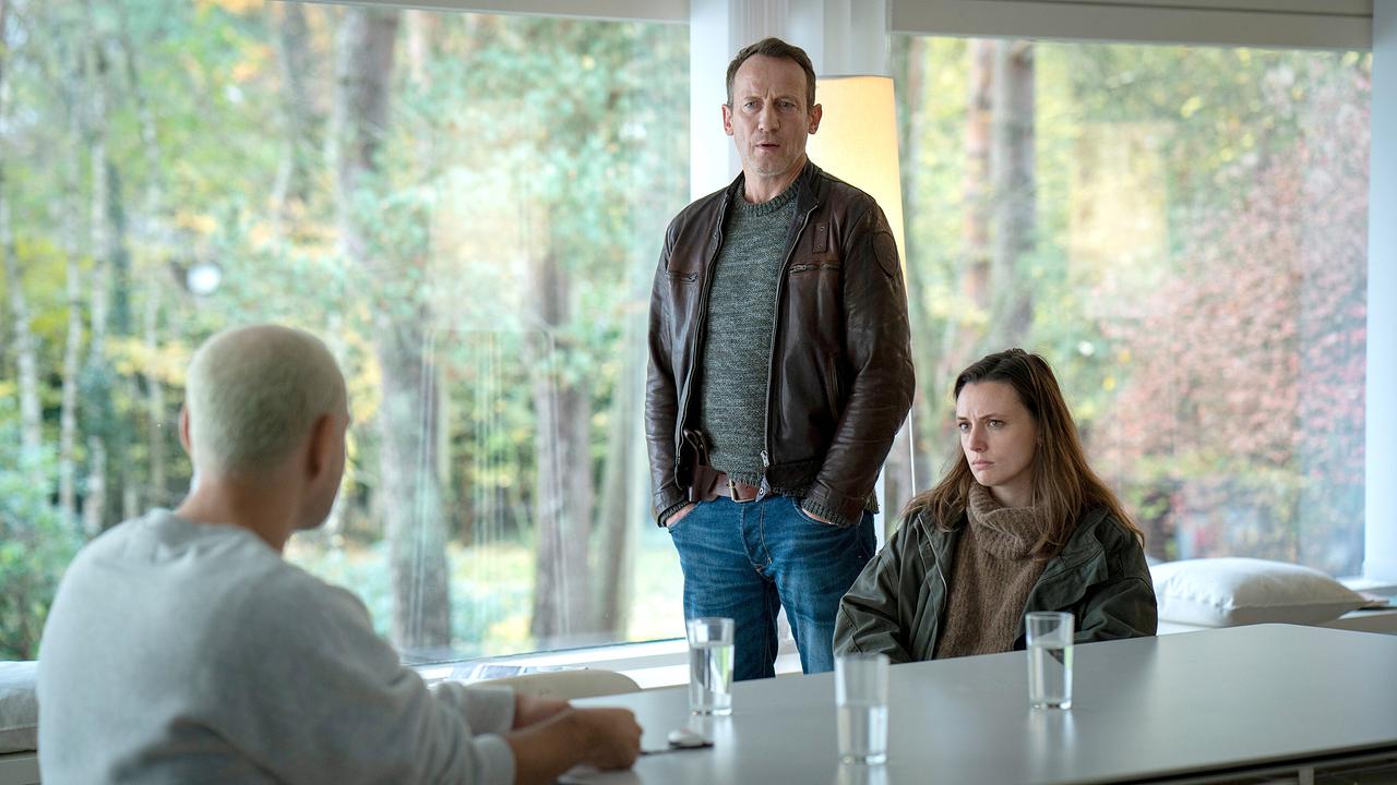 Falke und Lynn de Bear sprechen mit Carsten Kellmann mit blond gefärbten Haaren an einem Tisch mit Wassergläsern in einem modernen Wohnraum.