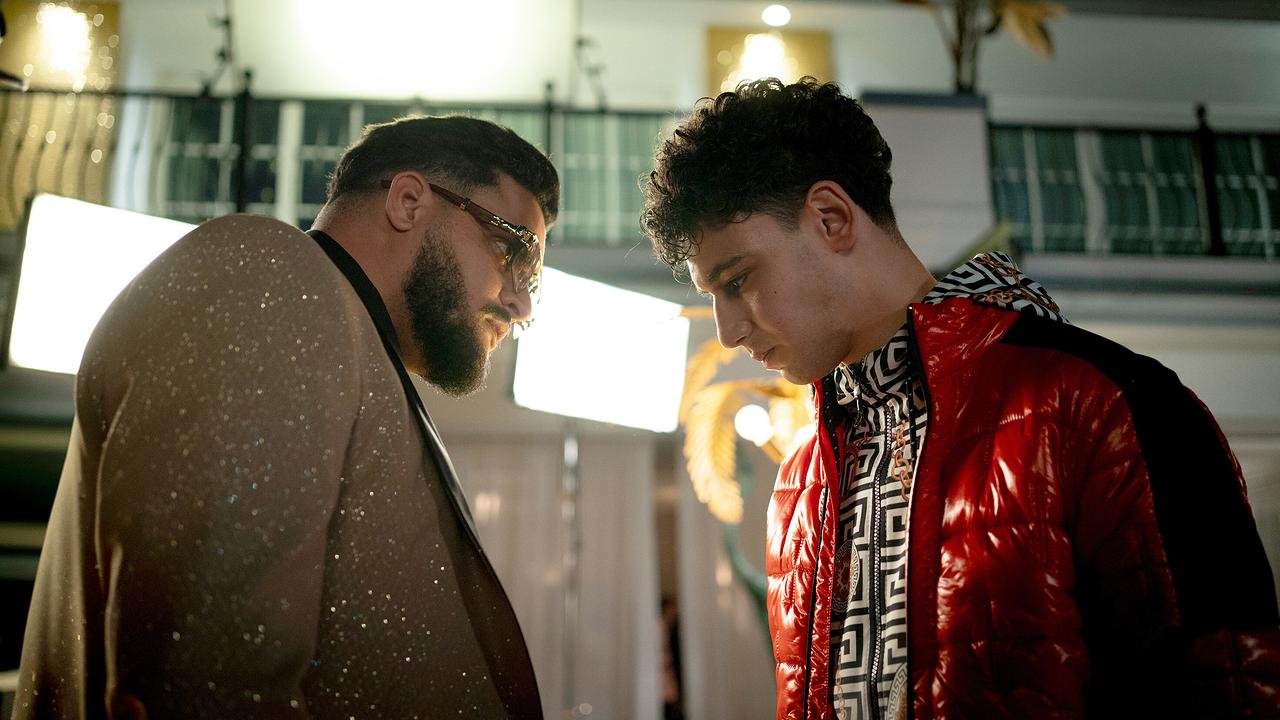 Sami im Glitzeranzug und Sonnenbrille im Gespräch mit dem zu Boden schauenden Karim Saidi in einer hell beleuchteten Umgebung