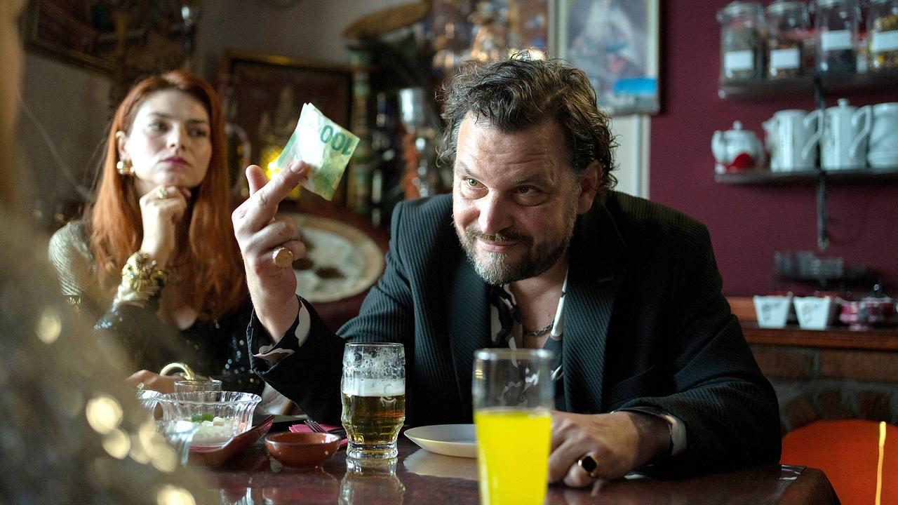 Dealer Ervin Zoric hält bei einem Gespräch mit jemandem an einem Tisch in einem Lokal einen 100-Euro-Schein in der Hand, während Frau Lulu daneben sitzt.