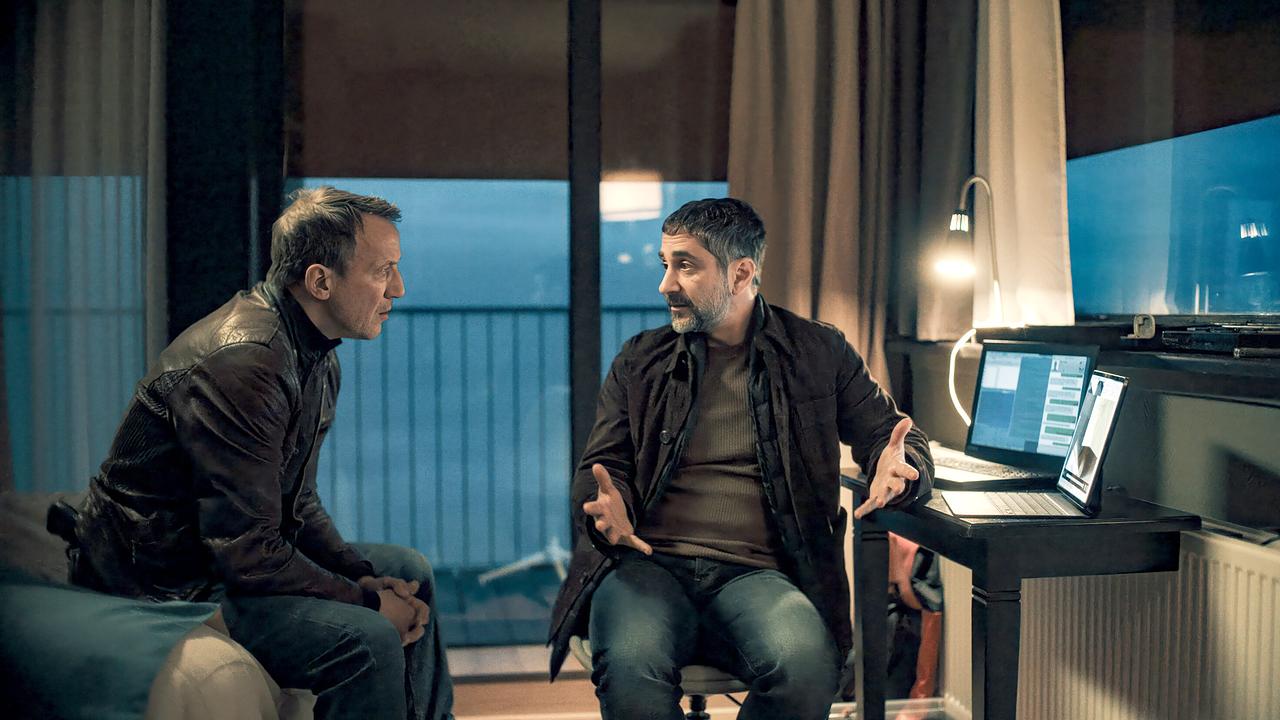 Falke und Mario Schmitt unterhalten sich in einem Raum mit einem geöffneten Laptop auf dem Tisch bei Abenddämmerung.