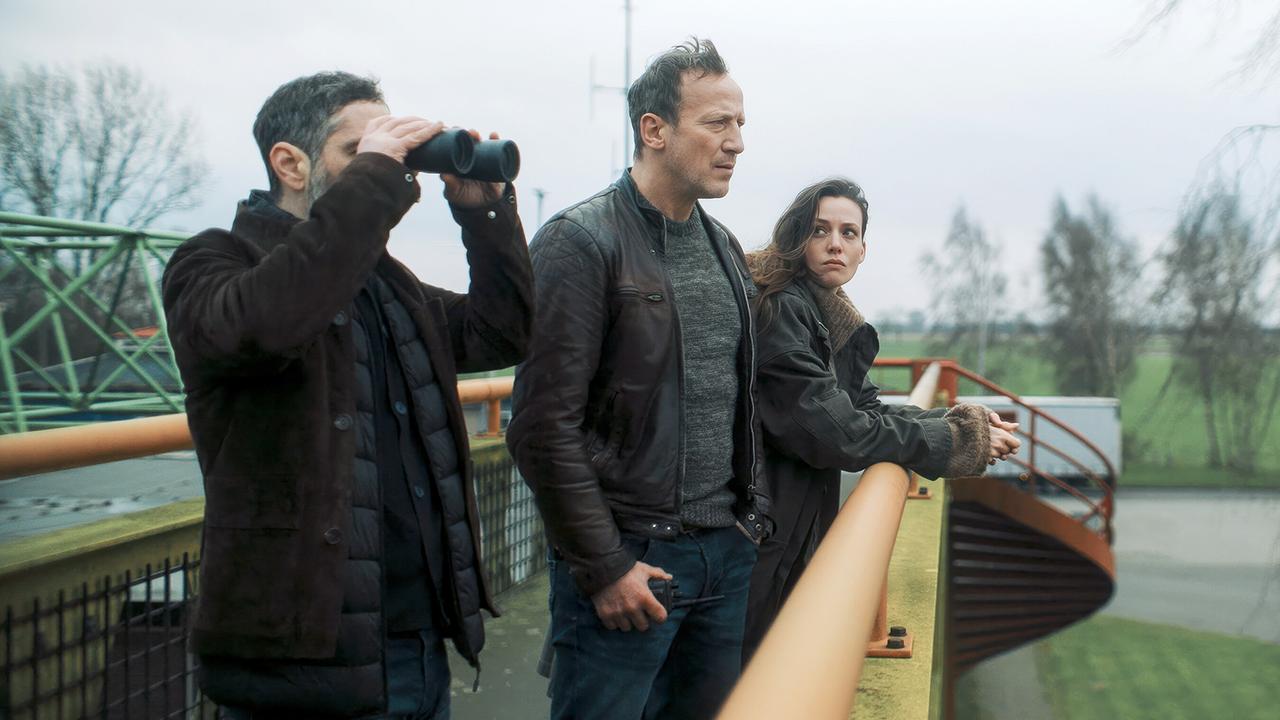 Mario Schmitt mit Fernglas, Falke  und Lynn de Baer stehen auf einer Brücke und beobachten etwas in der Ferne.