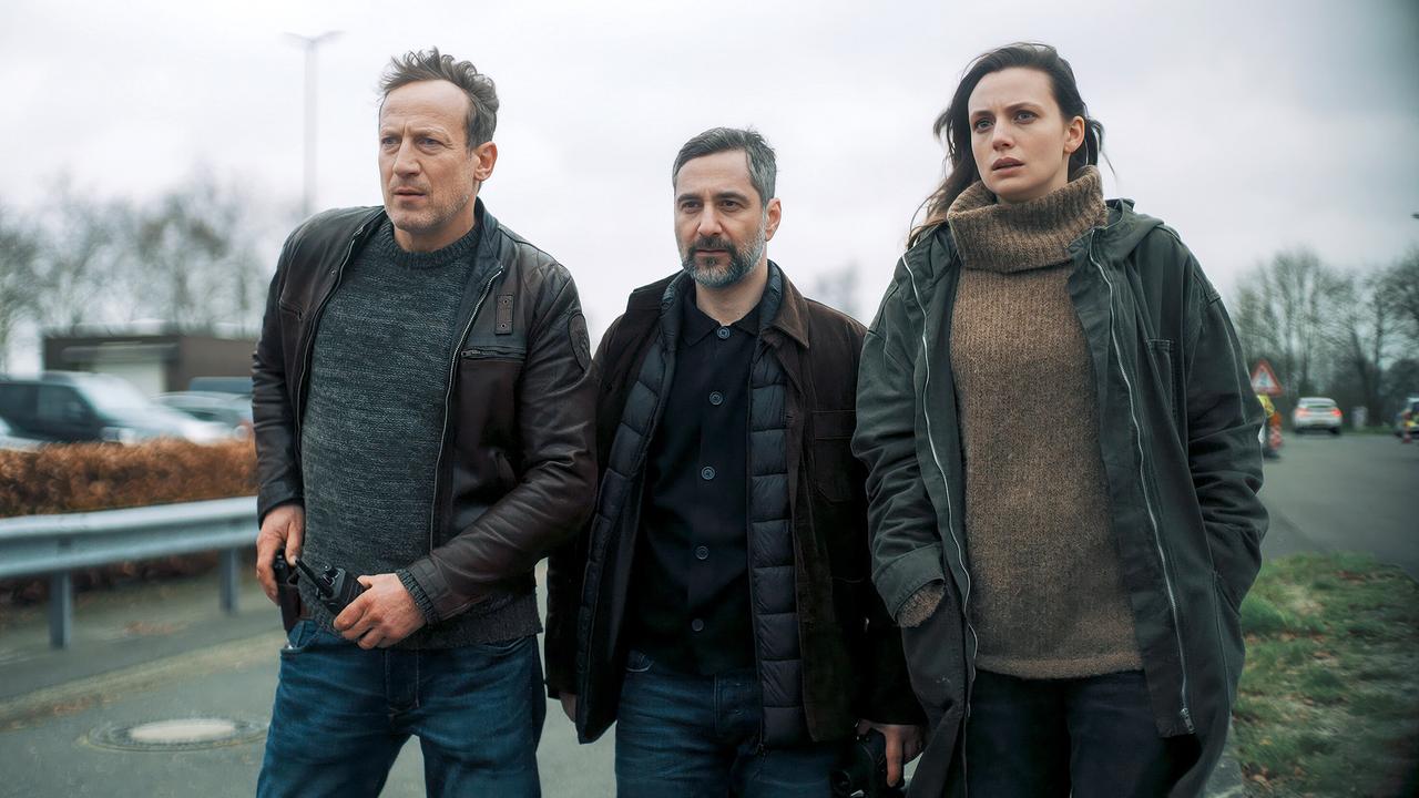 Falke, Mario und Lynn de Baer gehen nebeneinander eine Straße entlang, zwei von ihnen halten eine Waffe in der Hand.