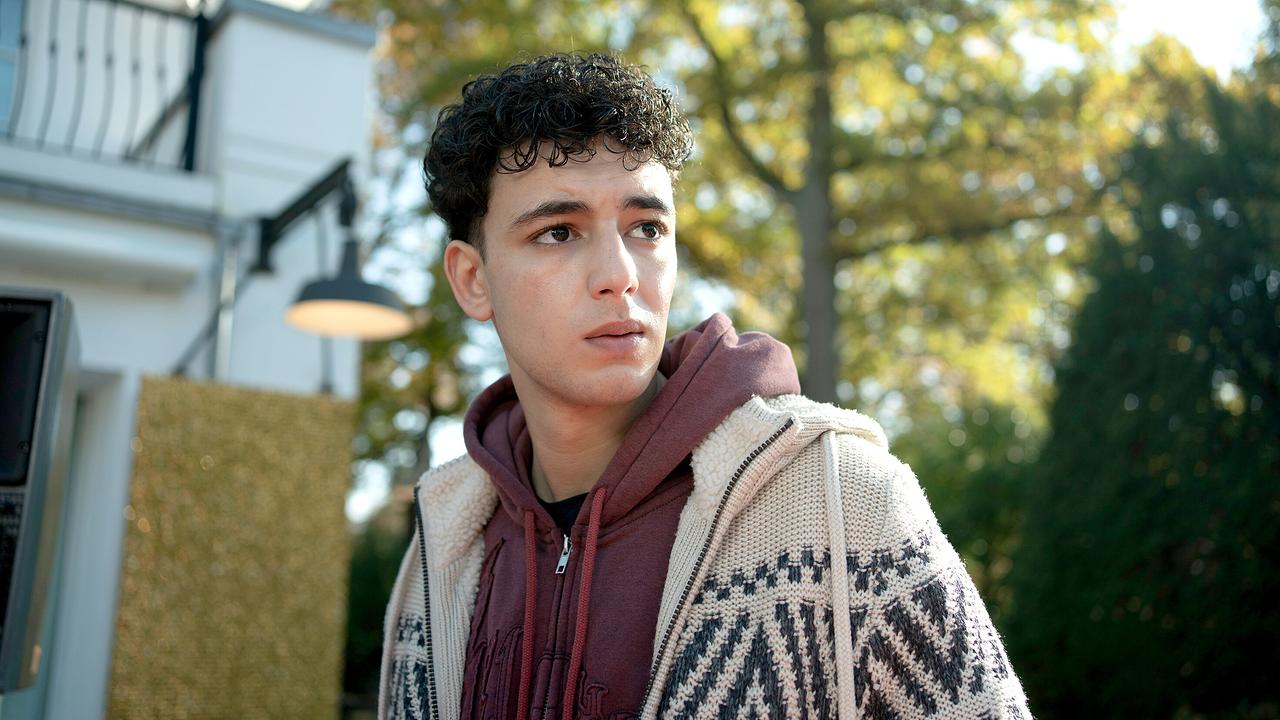 Sami in gemusterter Strickjacke und rotem Kapuzenpullover steht vor einem Haus im Freien bei Sonnenschein und blickt ernst.