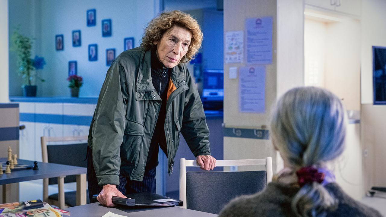 Bibi Fellner beugt sich über einen Tisch und spricht mit der älteren Anna in Strickjacke