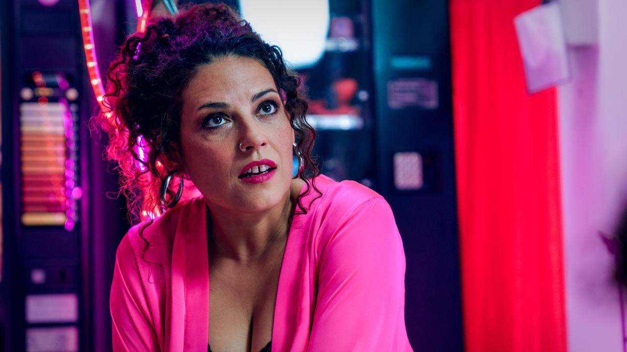 Ramona in pinkem Morgenmantel sitzt in einem Raum mit Neonlichtern und blickt zu jemandem auf.