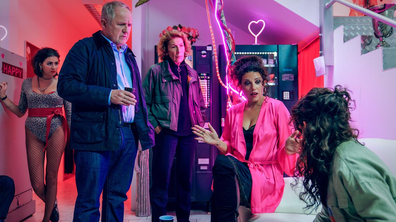 Moritz Eisner und Bibi Fellner und weitere Personen befinden sich in einem neonbeleuchteten Raum, während Ramona im pinken Morgenmantel auf einem Sofa sitzt und spricht.