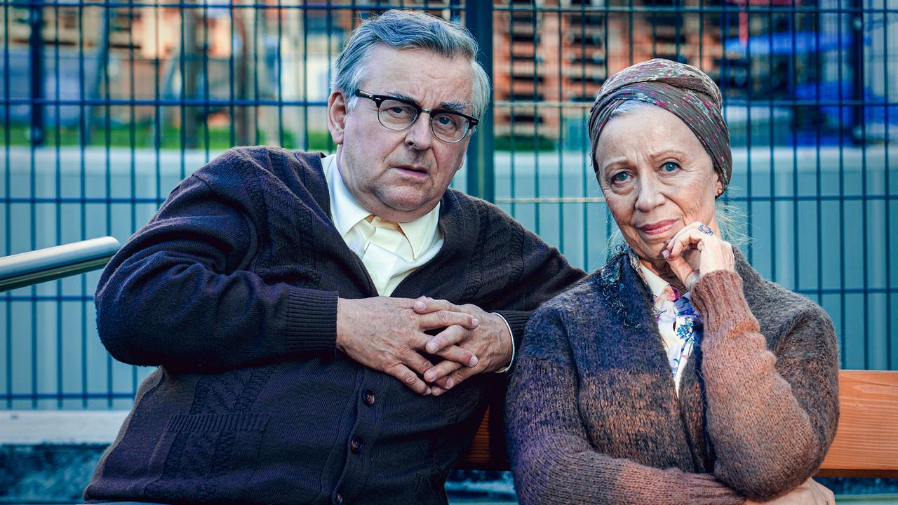 Fritz und Anna sitzen auf einer Bank im Freien vor einem Metallzaun.