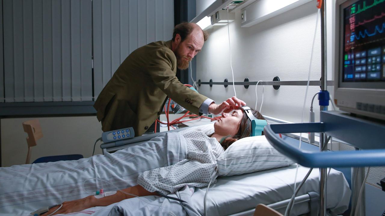 Dr. Schneider beugt sich im schwach beleuchteten Krankenzimmer über eine Patientenliege und berührt den Kopf der liegenden Patientin Eva Hütter.