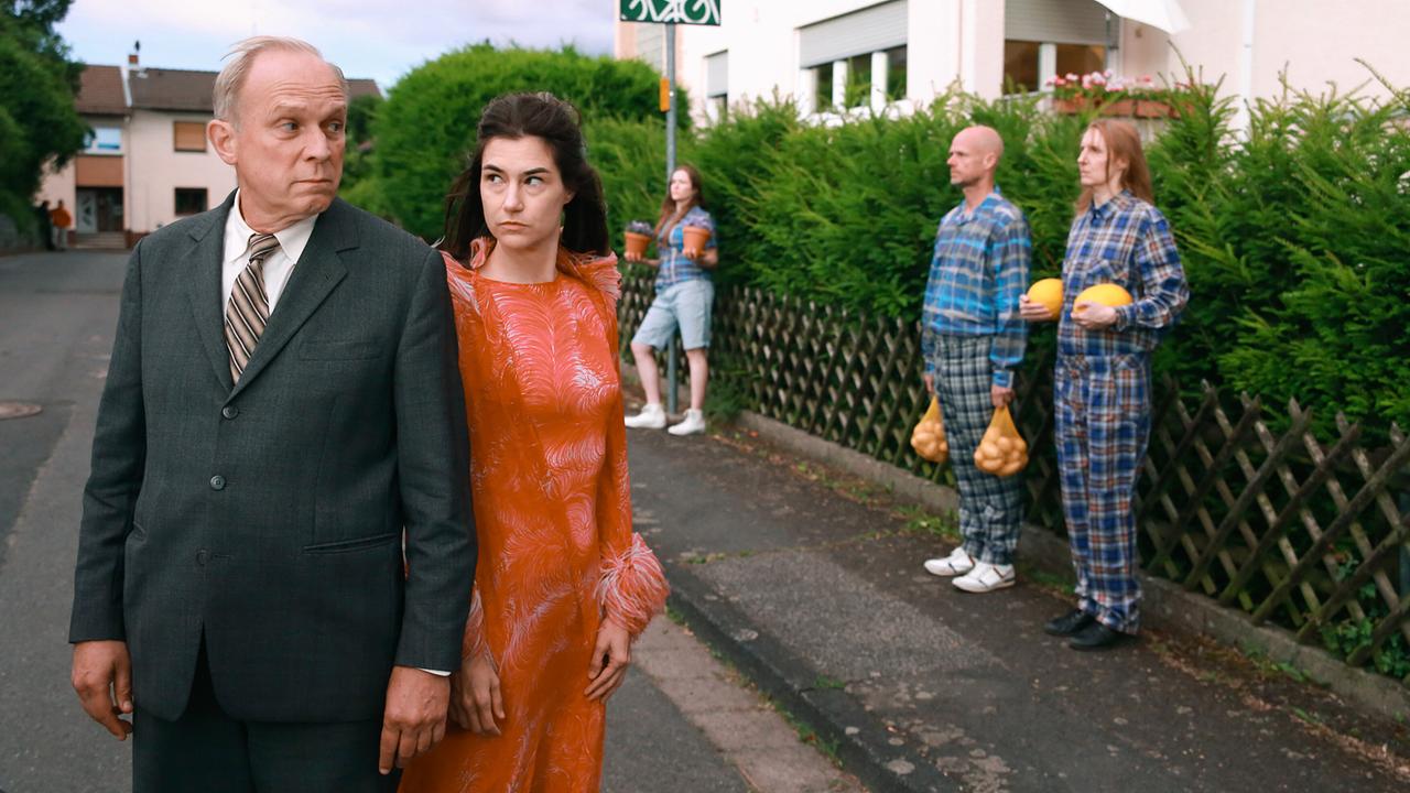 Felix Murot und Eva Hütter gehen auf einer Wohnstraße, während im Hintergrund ein Mann und eine Frau in karierten Anzügen und eine weitere Frau mit Melonen und Säcken oder Blumentöpfen posieren.