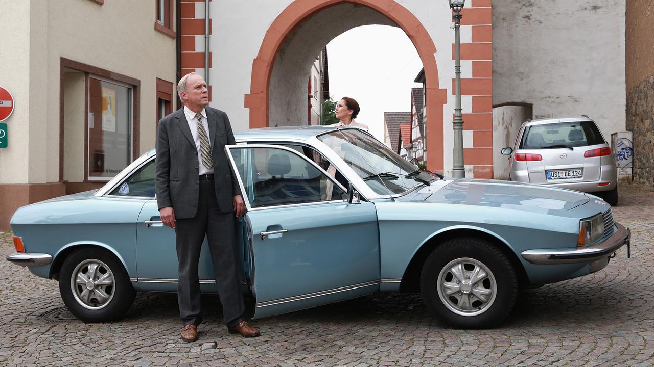Kommissar Felix Murot und Magda Wächter stehen neben einem hellblauen Oldtimer in einer Altstadt vor einem Torbogen.