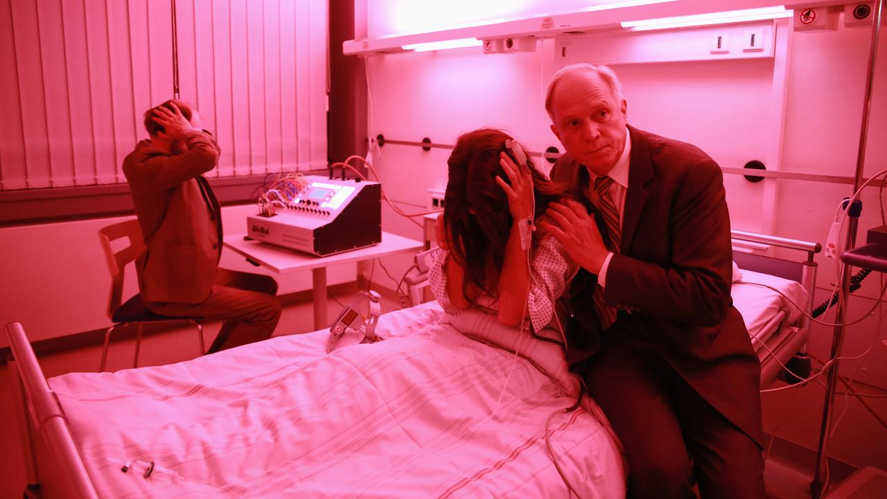 In einem pink-beleuchteten Krankenhauszimmer sitzt eine Patientin Eva Hütter verzweifelt im Krankenhausbett, während Felix Murot sie tröstet und Dr. Schneider im Hintergrund reagiert.