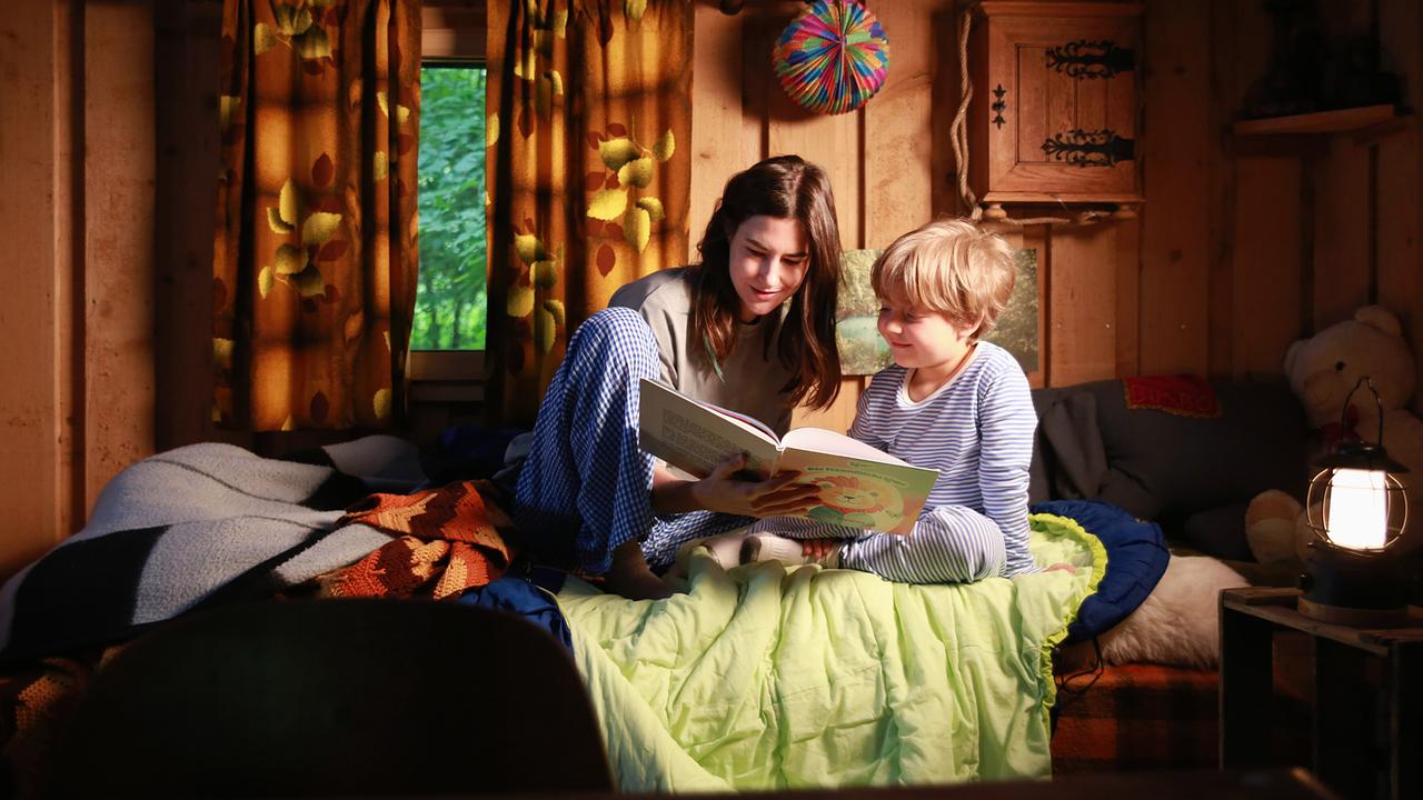 Eva Hütter liest Benjamin im Bett in einem gemütlichen Holzraum aus einem Buch vor.