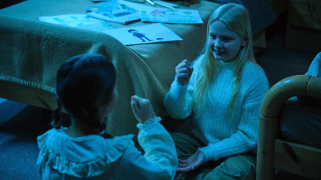 Eliza und ein weiteres Mädchen sitzen abends am Boden eines Zimmers und spielen miteinander, umgeben von Kinderzeichnungen.