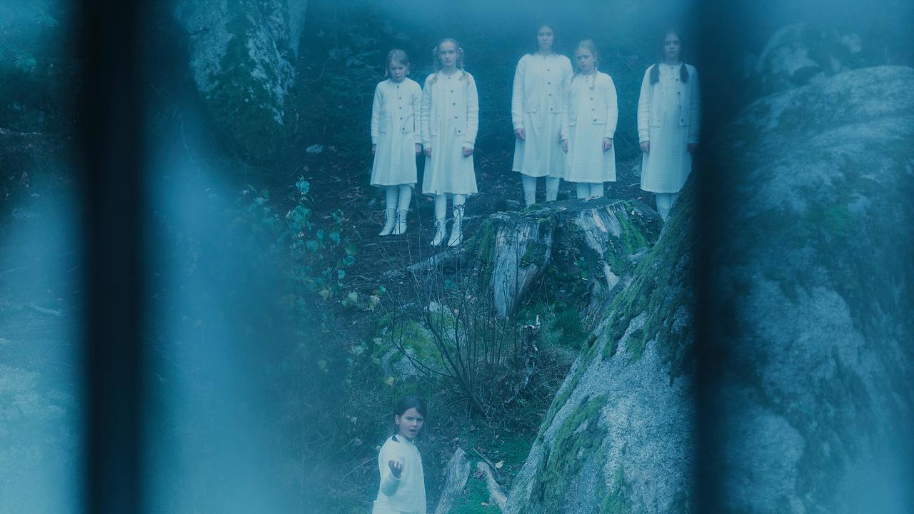 Eine Gruppe weiß gekleideter Kinder steht still im Wald, als würde sie auf jemanden warten.