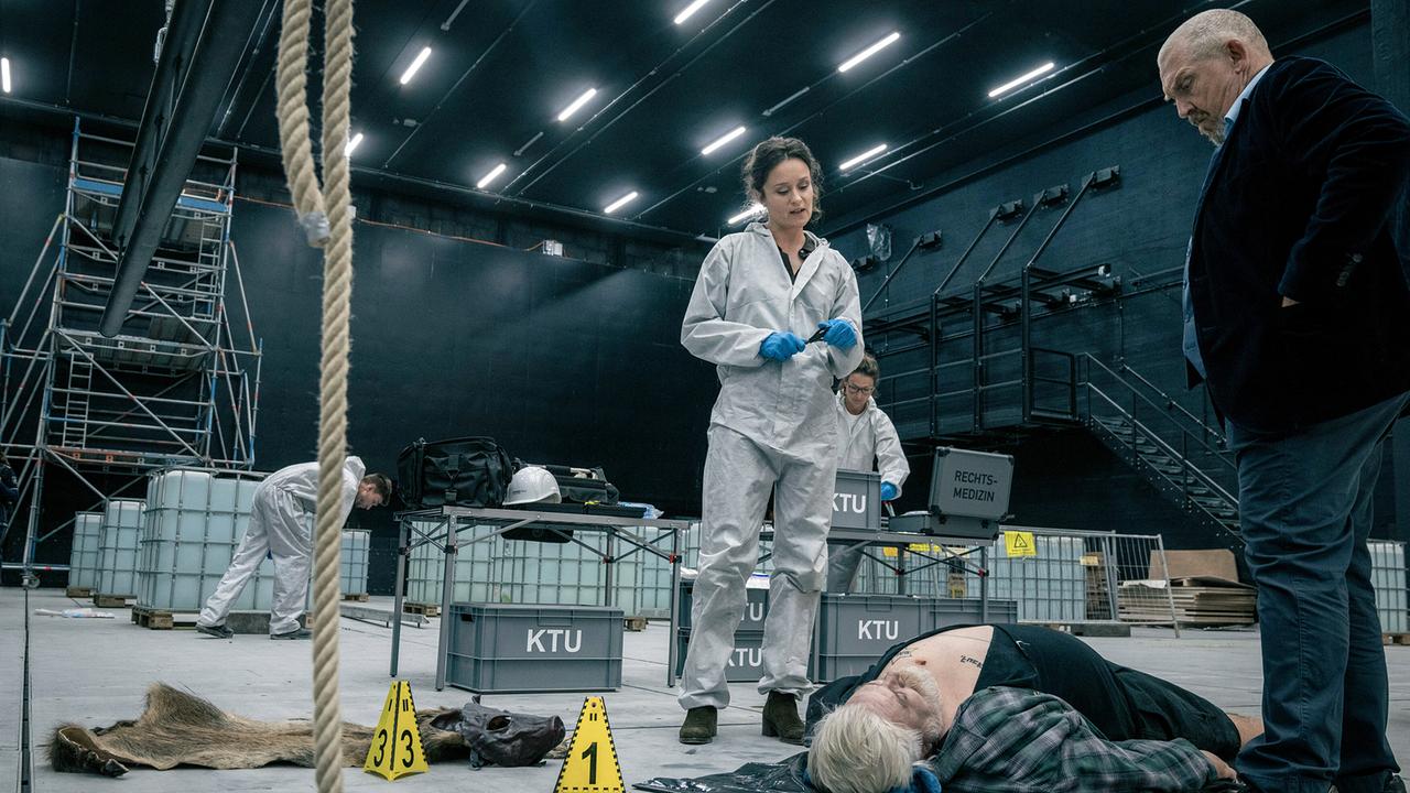 KTU-Leiterin Natalie Försten und Kommissar Freddy Schenk stehen in einem großen Bühnenraum am Tatort, an dem ein Toter am Boden liegt.