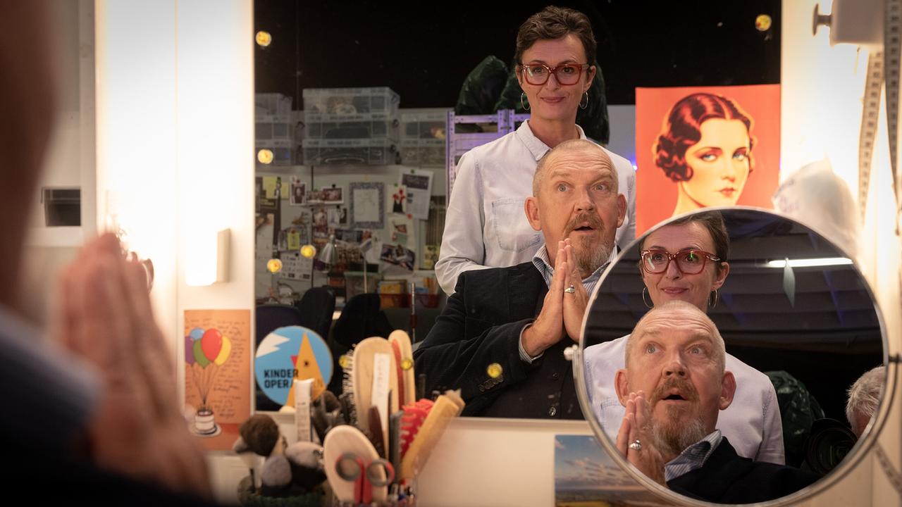 Maskenbildnerinnen Britt Maier steht hinter Kommissar Freddy Schenk vor einem beleuchteten Spiegel in der Maske einer Oper.