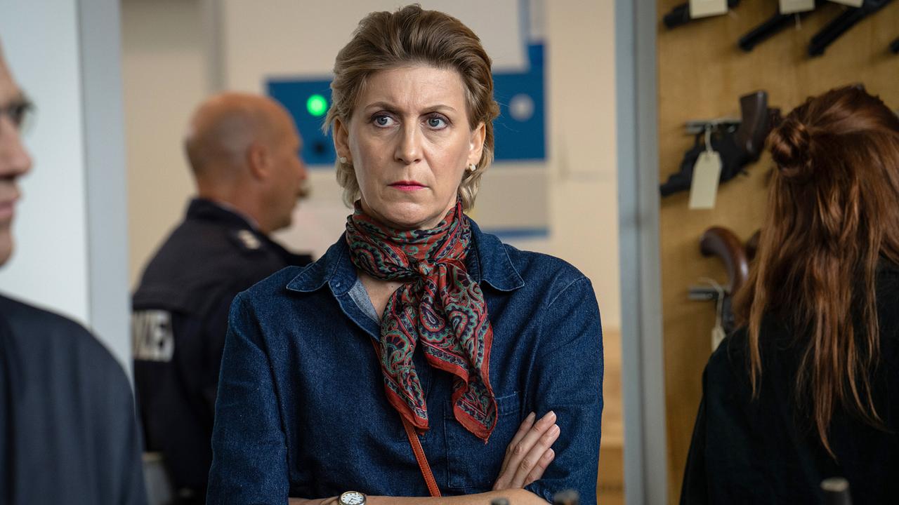 Operspielleiterin Jenny Ibsen steht mit verschränkten Armen in einem Raum, in dem an der Wand Pistolen hängen, während im Hintergrund Polizei zu sehen ist.
